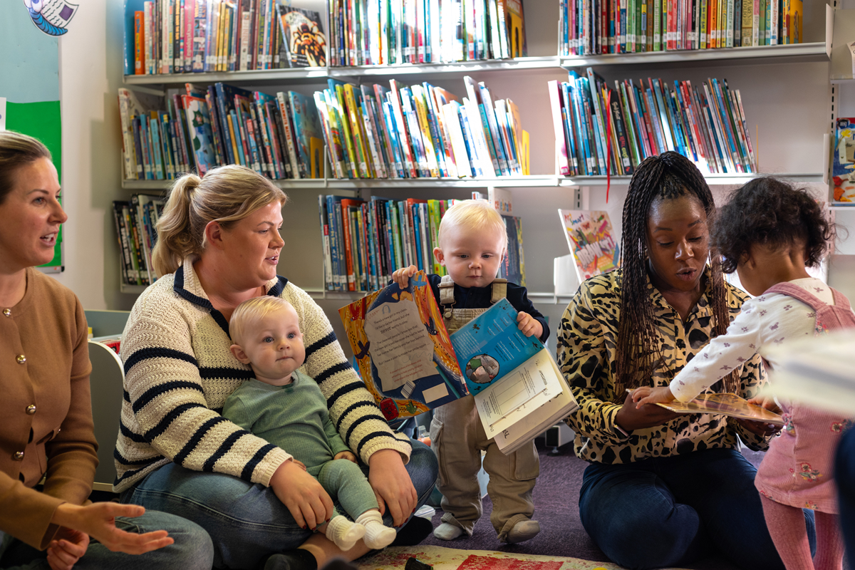 Adults and young children take part in a shared reading session in a library surrounded by bookshelves.